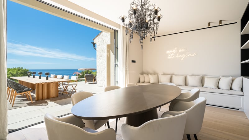 Salle à manger moderne avec table ovale sombre et chaises blanches ouvrant sur une terrasse ensoleillée surplombant la mer.