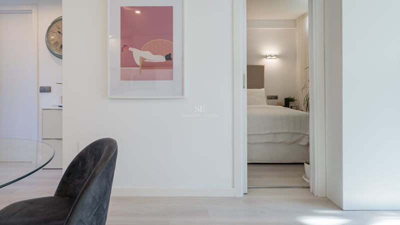 Vue à travers une porte ouverte sur une chambre minimaliste avec un grand lit, tête de lit beige et éclairage tamisé.