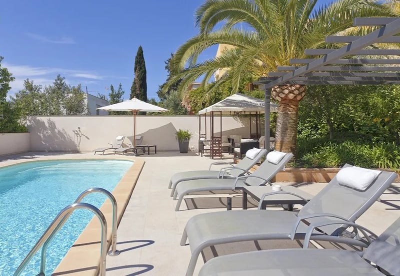 Piscine aux eaux turquoise entourée de transats gris, d'un palmier et d'une pergola sous un ciel bleu azur.