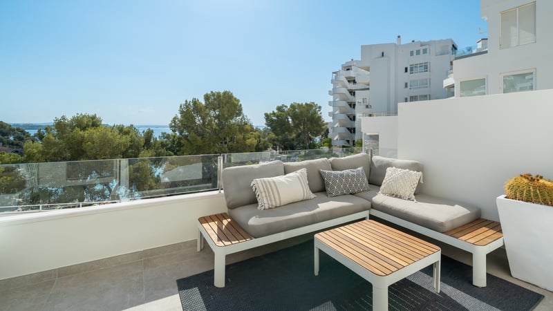 Terrasse sur le toit avec canapé gris en L, table en bois et garde-corps en verre surplombant des pins et la mer.