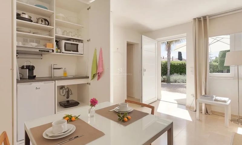 Kitchenette blanche moderne et table à manger avec porte ouverte sur un jardin ensoleillé et un palmier.