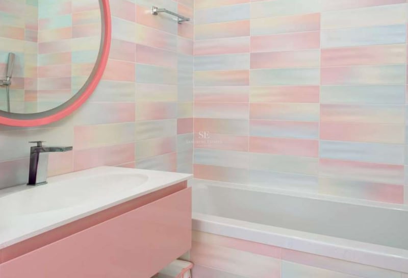 Salle de bains avec baignoire, meuble sous vasque rose et carrelage mural irisé aux couleurs douces.