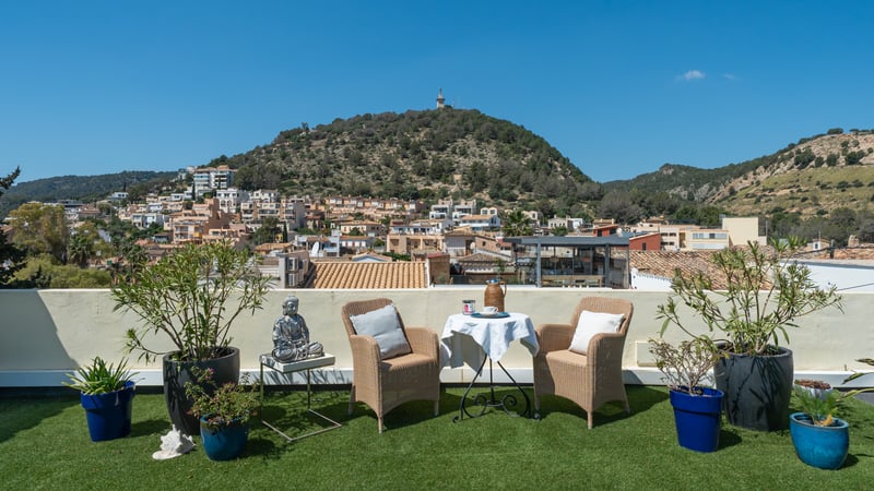 Terrasse sur le toit avec fauteuils en osier, table et plantes donnant sur un village et une colline méditerranéenne.