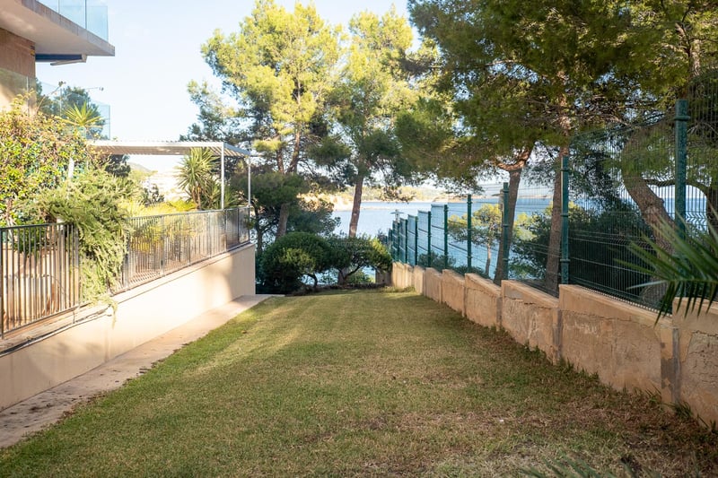 Une pelouse soignée entourée de pins et d'une clôture métallique, surplombant la mer Méditerranée turquoise.