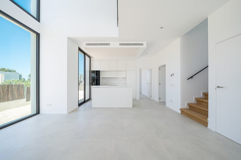 Cuisine blanche minimaliste avec îlot, escalier en bois et grandes baies vitrées du sol au plafond.