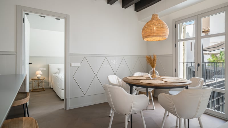Comedor moderno con mesa redonda de madera, sillas blancas y lámpara de mimbre junto a la entrada del dormitorio.
