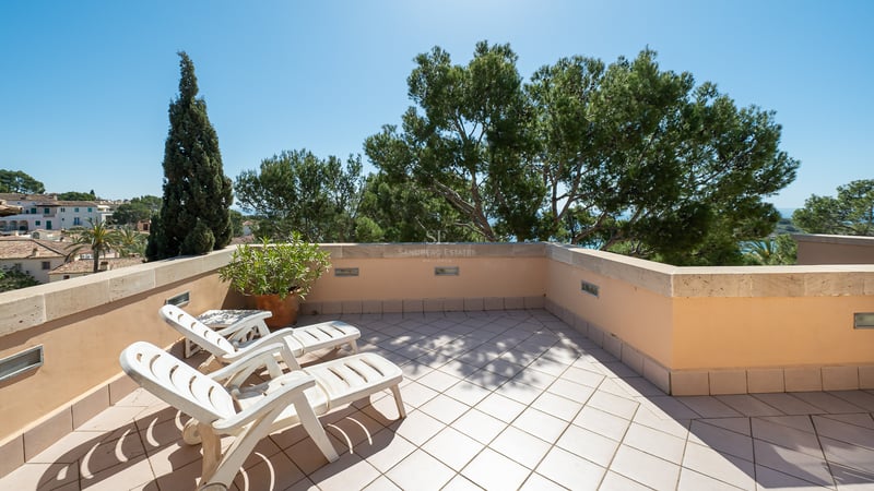 Terrasse de toit avec deux chaises longues blanches, carrelage en terre cuite et murs en pierre entourés de pins.