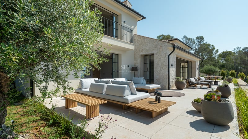 Salon d'extérieur contemporain sur terrasse en pierre avec olivier, mobilier en bois et façade de la villa.