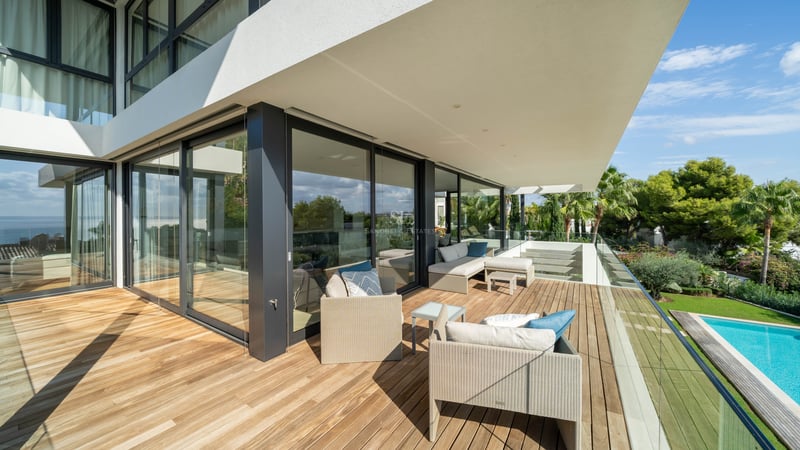 Terrasse spacieuse en bois avec mobilier lounge et garde-corps en verre surplombant une piscine et un jardin.