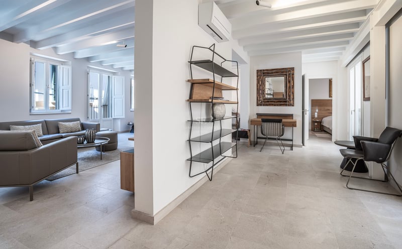 Grand salon moderne avec canapé en cuir gris, sols en pierre, poutres apparentes et étagère minimaliste.