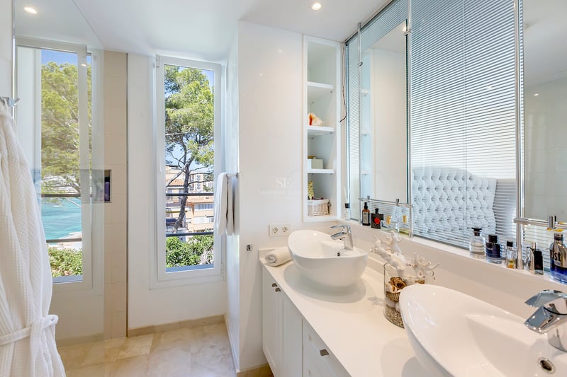 Salle de bain lumineuse avec deux vasques à poser blanches et fenêtre donnant sur la mer et des pins.