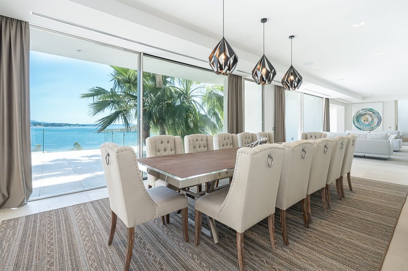 Salle à manger élégante avec une longue table, des chaises capitonnées beiges et de grandes baies vitrées sur la mer.