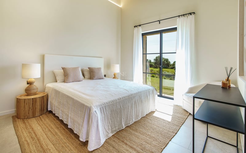 Dormitorio luminoso con cama blanca, alfombra de yute y ventanales con vistas a un entorno verde.
