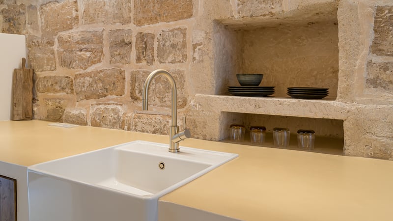 Évier blanc avec robinet doré devant un mur en pierre apparente avec des étagères intégrées dans une cuisine rustique.