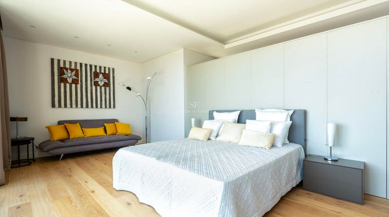 Chambre moderne lumineuse avec grand lit, parquet et canapé gris aux coussins jaunes.