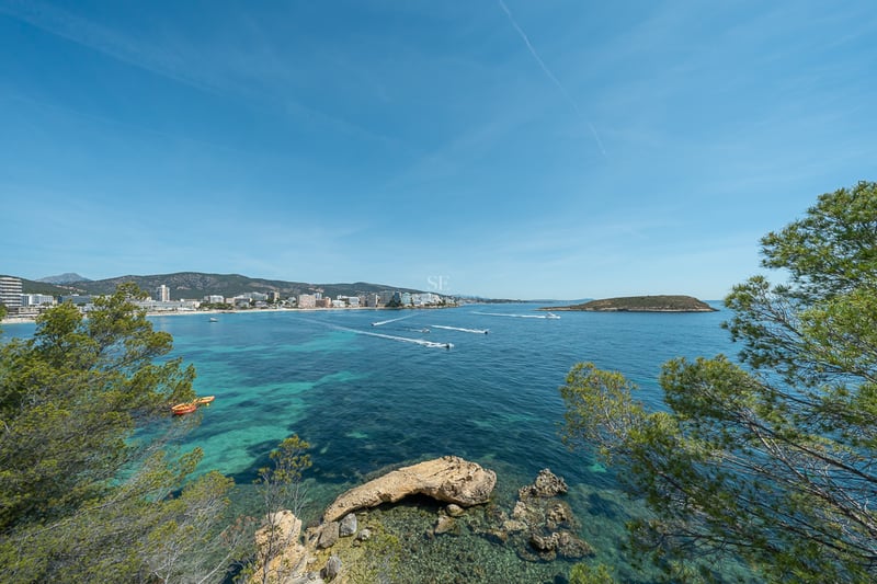 Vue d'une baie aux eaux turquoises avec des bateaux, entourée de pins et d'une ville côtière au loin.