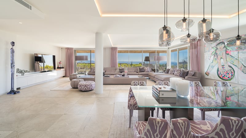 Salon ouvert moderne avec canapé d'angle mauve, table de salle à manger en verre et grandes baies vitrées sur la mer.