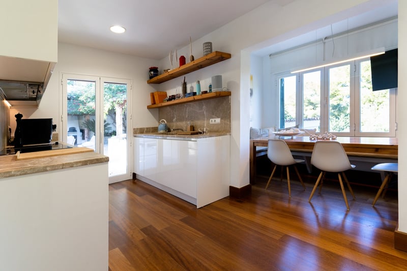 Cuisine blanche moderne avec plans de travail en pierre, étagères en bois et coin repas près de la fenêtre.