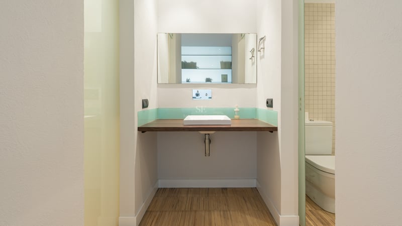 Salle de bain avec plan de travail flottant en bois, vasque carrée blanche, miroir et crédence en verre turquoise.