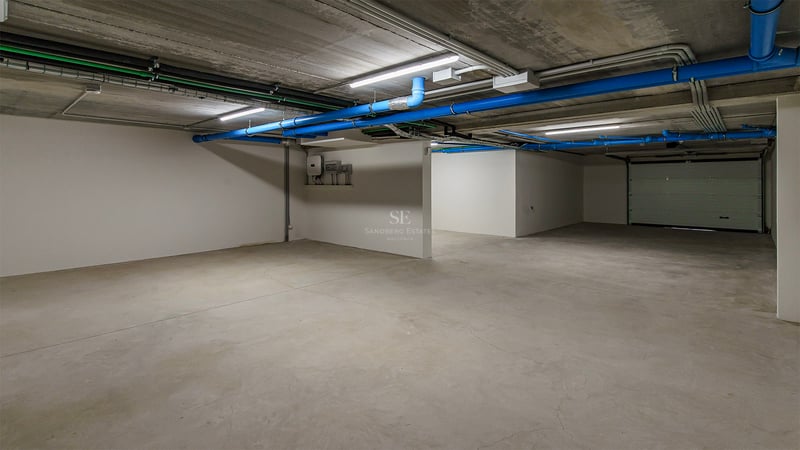 Garage souterrain vide avec sols en béton, tuyaux bleus et murs blancs sous éclairage LED.