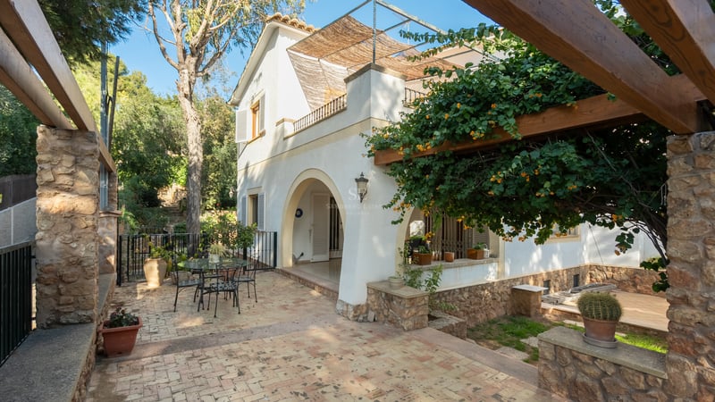 Patio ensoleillé en dalles, pergola en bois fleurie et façade de villa blanche avec arches.