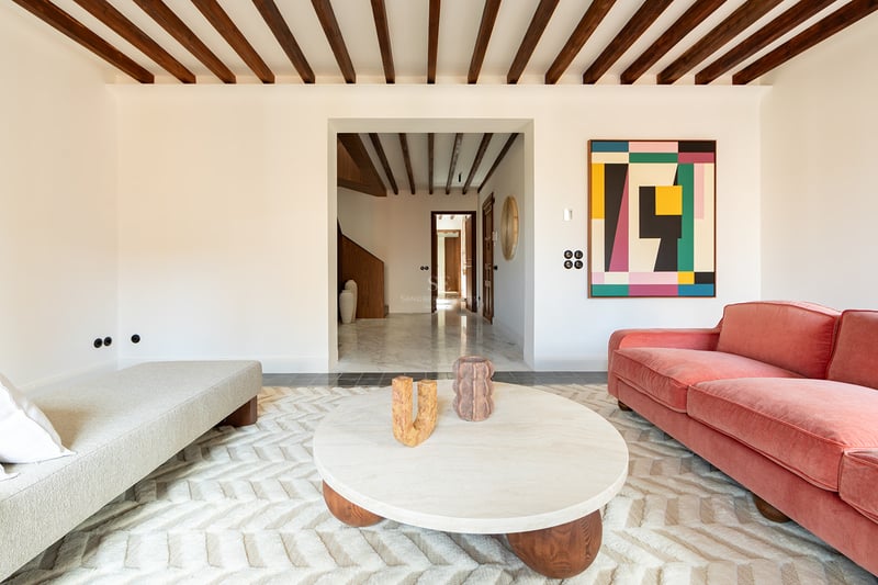 Salón moderno con vigas de madera oscura, sofá color terracota, mesa de mármol redonda y alfombra con textura.