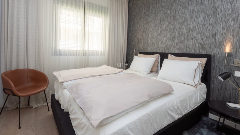 Chambre moderne avec lit sombre, mur texturé gris, fauteuil en cuir tabac et rideaux blancs toute hauteur.