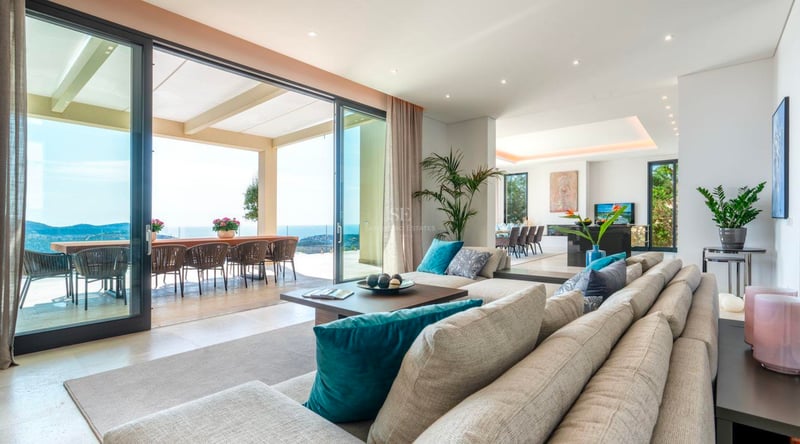 Vaste salon moderne avec canapé beige, baies vitrées toute hauteur et terrasse donnant sur la mer.