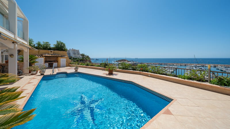 Piscine ensoleillée sur une terrasse en terre cuite surplombant une marina méditerranéenne et la mer bleue.