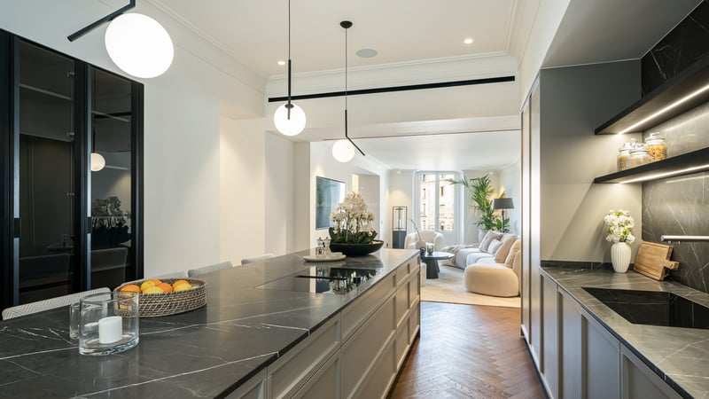 Modern kitchen featuring a dark marble island and herringbone wood floors, opening into a bright living room area.
