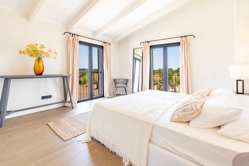 Bright master bedroom featuring white vaulted ceilings with exposed beams, two sets of dark-framed French doors, and oak flooring.