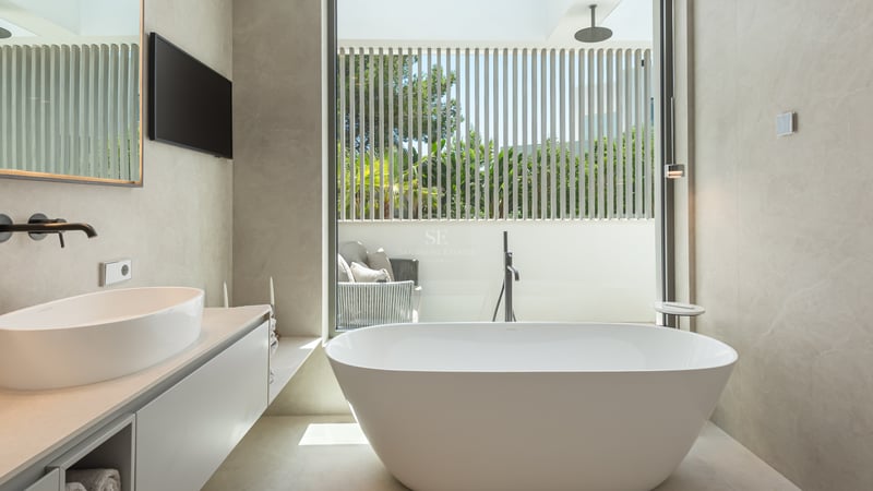 Salle de bains minimaliste avec baignoire îlot blanche, vasque à poser et grande fenêtre sur terrasse privée ensoleillée.