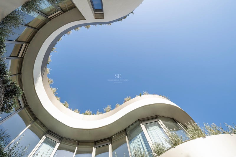 Vue en contre-plongée d'une façade blanche courbée avec de grandes baies vitrées et de la végétation sur le toit.