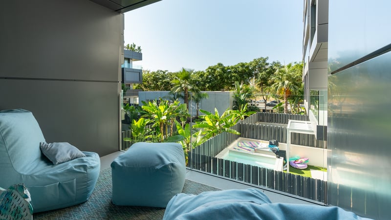 Balcon moderne avec poufs bleu clair et garde-corps en verre surplombant un jardin tropical et une piscine.