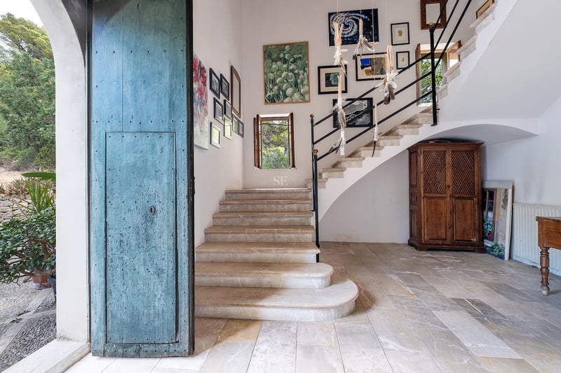 Hall d'entrée avec une grande porte bleue vieillie, escalier en pierre et galerie de tableaux sur murs blancs.