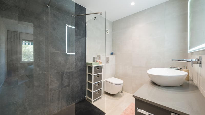 Salle de bain moderne avec douche en verre, carrelage gris foncé, vasque blanche et toilettes suspendues.