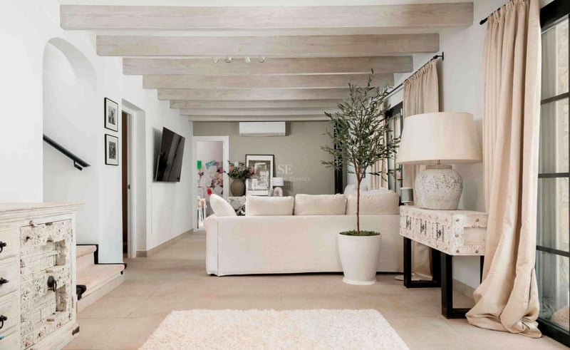 Salon lumineux avec canapé blanc, poutres en bois clair, sol en pierre et grande baie vitrée.