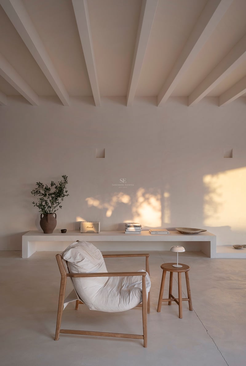 A minimalist room with white beamed ceilings, a wooden armchair, and warm golden hour light hitting a plastered wall.