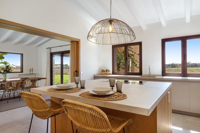 Îlot de cuisine moderne avec tabourets en osier et plafond à poutres blanches avec vue sur la campagne.