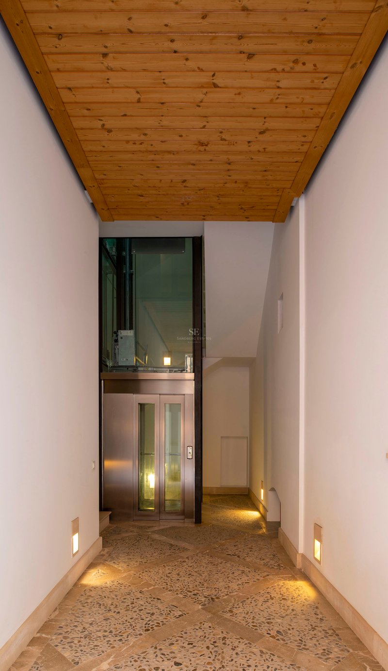 Couloir moderne avec sol en mosaïque de pierre, plafond en bois et ascenseur en verre au fond.