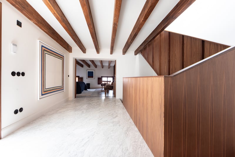 Pasillo luminoso con suelos de mármol blanco, vigas de madera oscura en el techo y una escalera moderna de madera.