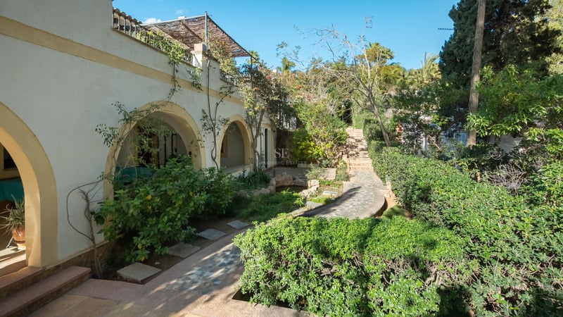Sentier en pierre à travers un jardin méditerranéen luxuriant à côté d'une villa blanche aux arcades.