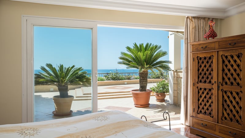 Chambre principale lumineuse avec armoire en bois et baies vitrées ouvrant sur une terrasse avec palmiers et vue mer.