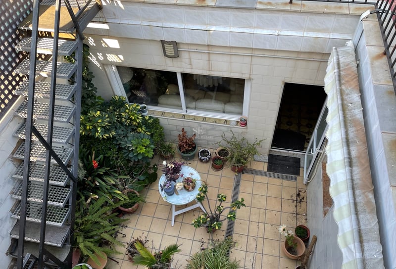 Vue d'en haut d'une terrasse carrelée avec escalier métallique, nombreuses plantes en pots et petite table ronde.