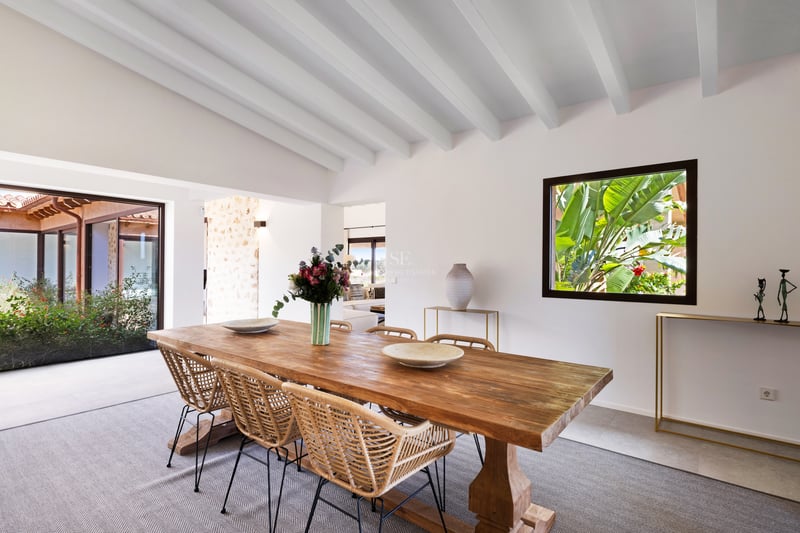 Salle à manger lumineuse avec table en bois, chaises en osier et plafond à poutres blanches.