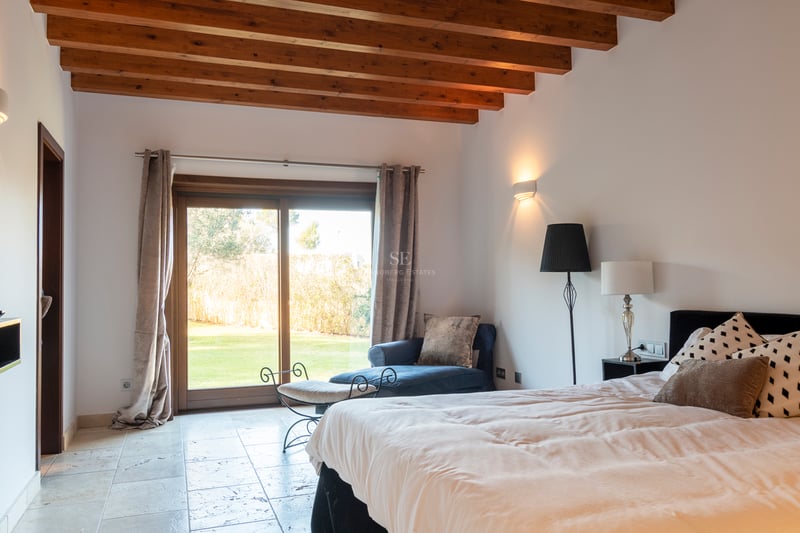 Chambre spacieuse avec poutres apparentes en bois, sol en pierre et baie vitrée ouvrant sur un jardin.