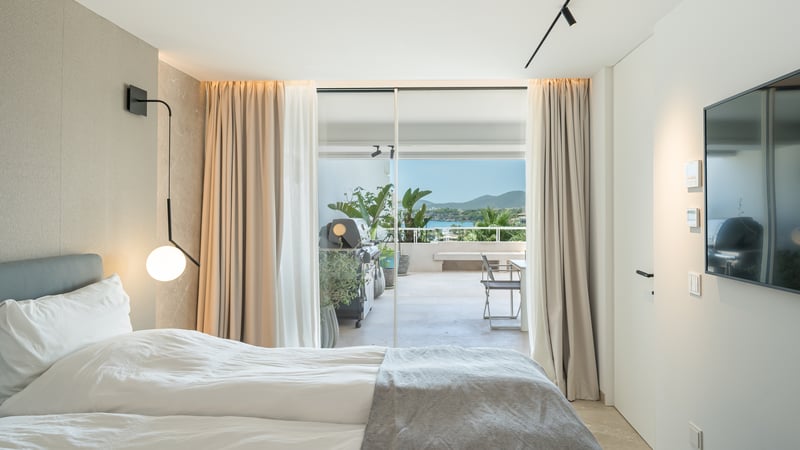 Chambre moderne avec lit blanc et portes vitrées s'ouvrant sur une terrasse avec barbecue et vue sur la mer.