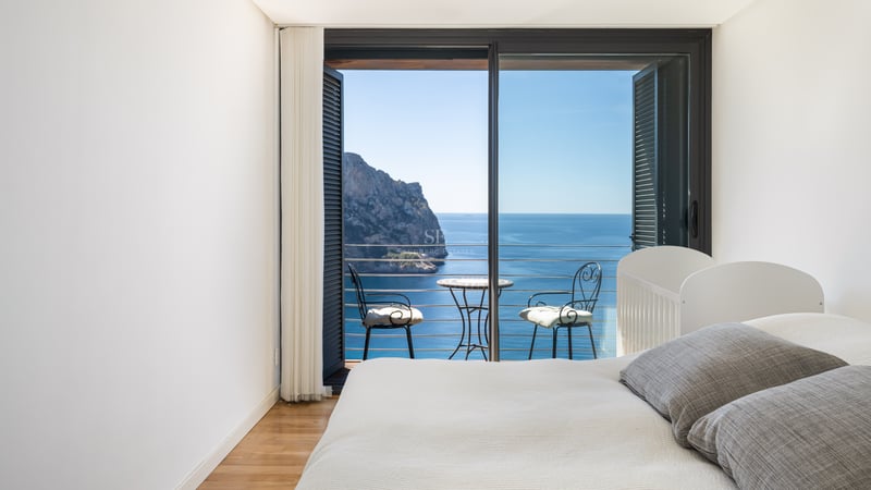 Chambre minimaliste blanche avec balcon privé, mobilier en fer forgé et vue sur une falaise et la mer Méditerranée.