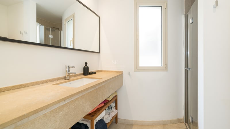 Salle de bain moderne avec lavabo en pierre beige, grand miroir et douche vitrée dans une pièce lumineuse.