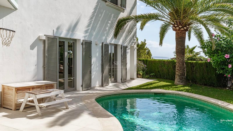Piscine turquoise à côté d'une villa blanche avec un palmier, des volets gris et une vue lointaine sur l'océan.